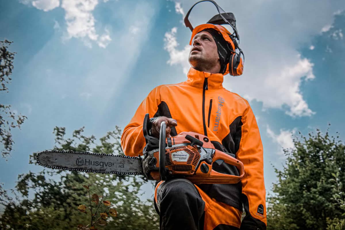 Arborist i orange skyddsjacka och hörselskydd med motorsågen i händerna mot en ljus himmel.