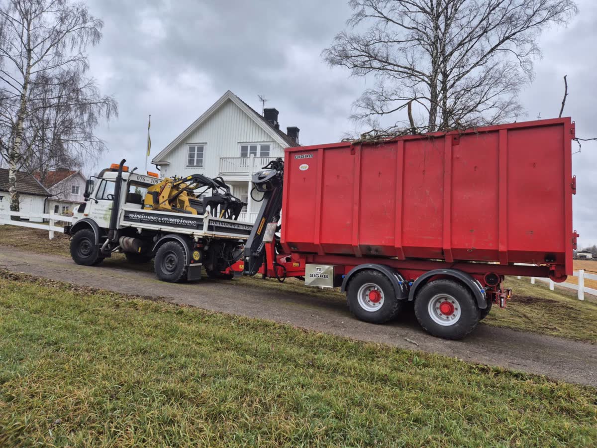 Lastbil med röd container och kranmaskin på uppfarten vid ett vitt hus.